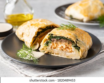 Salmon Wellington with spinach and mushrooms (champignons) baked in puff pastry served with creamy sauce and green dill on black ceramic plate. Homemade tasty pie. Traditional fish dish. Close up view