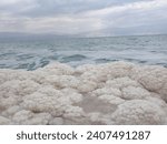 Salt formations on the shore of the Dead Sea with mountains and overcast sky in the background.