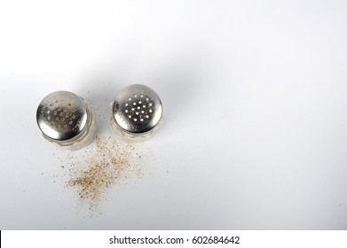 Salt And Pepper Shakers Shot From Above On White Surface. Horizontal Photo With Copy Space In The Right Side.  Salt And Pepper Grains On The Surface.