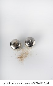 Salt And Pepper Shakers Shot From Above On White Surface. Vertical Photo With Copy Space In Upper Part.  Salt And Pepper Grains On The Surface.