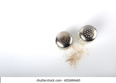 Salt And Pepper Shakers Shot From Above On White Surface. Horizontal Photo With Copy Space In The Left Side.  Salt And Pepper Grains On The Surface.