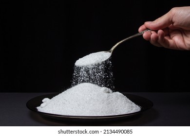 Salt is poured from a spoon onto a plate with salt on a black background. Excessive salt intake. Coarse Rock salt