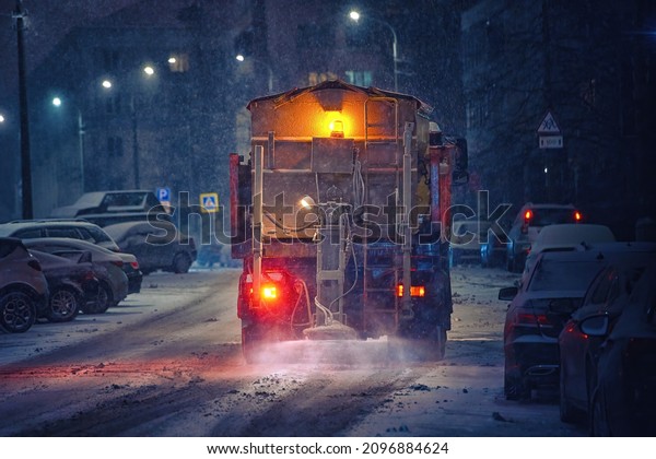 Salt spreading. Snow plow service truck removing\
snow and spreading salt on snowy city road during blizzard, night\
work road maintenance. Truck spreading de-icing salt on snowy and\
icy asphalt road