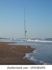 Salt Water Fishing Rods In The Sand.  Waves Approaching The Shore With White Foam And Blue Sky Overhead.  