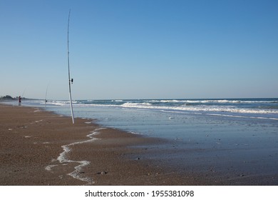 Salt Water Fishing Rods In The Sand.  Waves Approaching The Shore With White Foam And Blue Sky Overhead.  