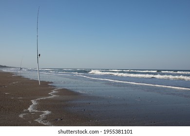 Salt Water Fishing Rods In The Sand.  Waves Approaching The Shore With White Foam And Blue Sky Overhead.  