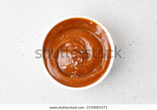 Salted caramel sauce in a\
white saucer on grey background. Curl of caramel with sea salt\
pieces. Top view.
