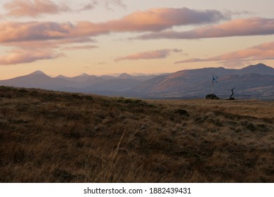 Saltire Flying On Top Of A Hill With Mountains In The Background Under A Red Sunset Sky With Fluffy Clouds