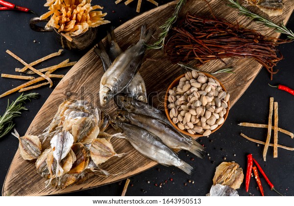 Salty dried fish roach and yellow stripe scad fish\
with dried fish sticks and pistachios on wooden board on black\
table background. Top\
view.