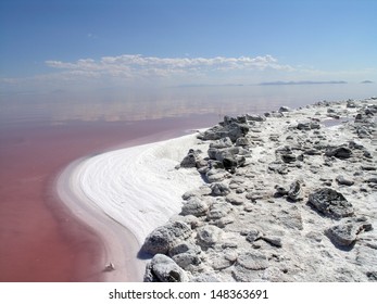 The Salty Shore Of The Great Salt Lake With Mountains In The Background. The Water Looks Pink Due To A Special Algae That Grows In High Levels Of Salt.  Clouds Float In The Sky.