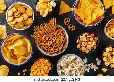 Salty snacks, party mix. An assortment of crispy appetizers, overhead flat lay shot on a black background. Potato and tortilla chips, crackers, popcorn etc
