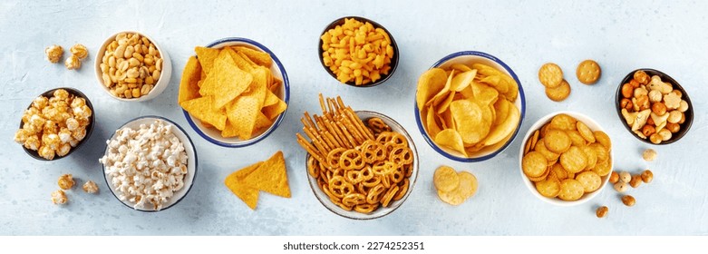 Salty snacks, party mix, overhead flat lay panorama. An assortment of appetizers. Potato and tortilla chips, crackers, popcorn etc panoramic banner
