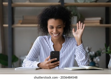 Salut. Smiling Biracial Female Teenager Sit At Workplace Take Break In Work Study To Make Or Answer Video Call On Phone. Happy Young Black Lady Wave Hand Hi Contact Friend In Videochat Conference App