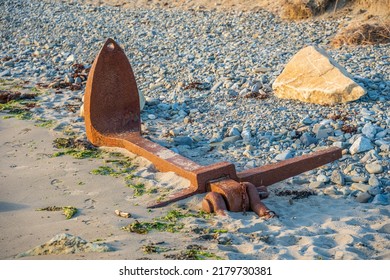 Salvaged Rusty Shipwrecked Boats Anchor From The Sea Bed  Partially Buried In The Sand On A Rocky Pebble Beach