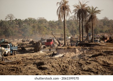 Sambouya, Gambia, Africa, December 15, 2020, horizontal wide angle photography: landscape with sandy ground, many holes, palm trees and sand mining big trucks, with bright sky, outdoors on a sunny day