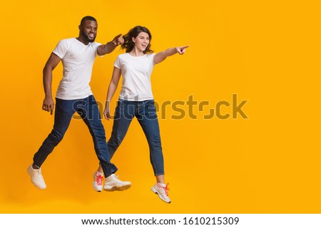 In the same direction. Jumping interracial couple pointing at copy space over yellow background, full length shot