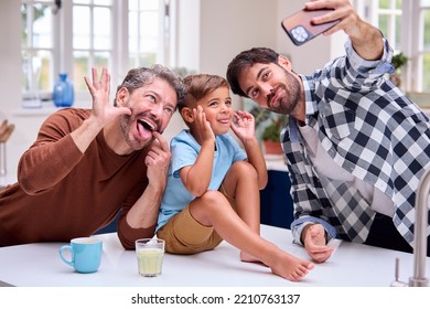 Same Sex Family With Two Dads Pulling Faces For Selfie In Kitchen With Son Sitting On Counter