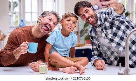 Same Sex Family With Two Dads Pulling Faces For Selfie In Kitchen With Son Sitting On Counter