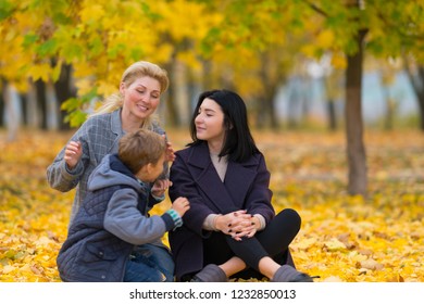 A Same Sex Female Couple With Their Son In A Happy Family Portrait At A City Park With Yellow Fall Leaves.