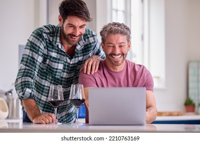 Same Sex Male Couple Drinking Wine Using Laptop At Home To Check Finances Book Holiday Or Shop