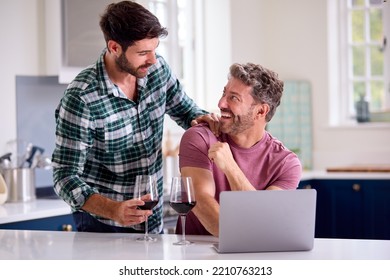 Same Sex Male Couple Drinking Wine Using Laptop At Home To Check Finances Book Holiday Or Shop