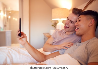 Same Sex Male Couple Lying In Bed At Home Pulling Faces For Selfie On Mobile Phone