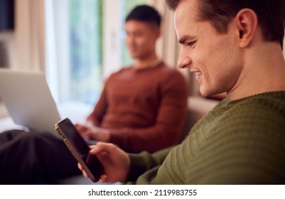 Same Sex Male Couple Sitting On Sofa At Home Using Mobile Phone And Laptop