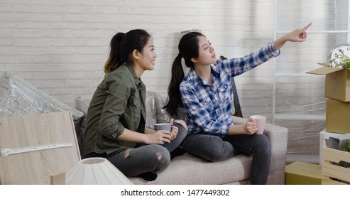 Same Sex Marriage People Mortgage And Real Estate Concept. Happy Lesbian Couple Pointing Showing Window Looking From New Home Sitting On Couch Sofa In Living Room With Packing Parcel Boxes Cardboard