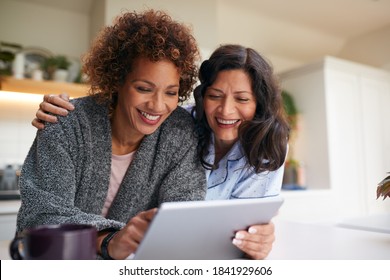 Same Sex Mature Female Couple At Home In Kitchen In Pyjamas Drinking Coffee With Digital Tablet