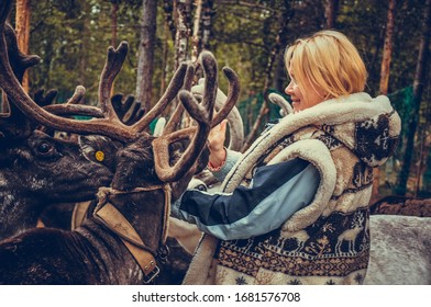 Sami Village, Sam-Syyt / Russia - August 6 2019: A Girl In Sami Clothes Stands Next To A Reindeer And Feeds Them