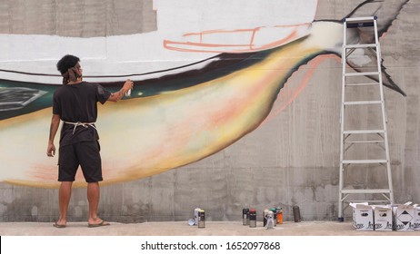 Samutsakhon, Thailand - February21,2020 : Rear View Of Asian Street Artist Is Painting Graffiti On Cement Wall With Ladder And Various Colors Aerosol Spray Cans On The Street In Public Area  