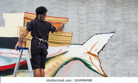 Samutsakhon, Thailand - February21,2020 : Rear View Of Asian Street Artist In Black Casual With Various Colors Aerosol Spray Cans On Ladder Is Painting Graffiti On Cement Wall In Public Area 