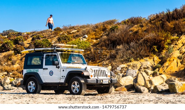 San Diego, California - October 08 2017: San Diego\
California. State Parks Life Guard off road truck parked on sea\
shore of west coast city beach on summer day. Shirtless man walking\
on cliff above.