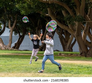 San Diego, California, USA - 5 June 2022: Children Play On The Grass In Tuna Harbor Park Chasing Soap Bubbles, Which Shine Irridescent Against Dark Trees. They Laugh, Jump And Have Fun Bursting Them.