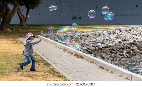 San Diego, California, USA - 5 June 2022: A Child Plays On The Shore Of Tuna Harbor Park Chasing Irridescent Soap Bubbles. The Kid, With Orange Sun Hat, Has Fun Running To Catch Them Before Bursting.