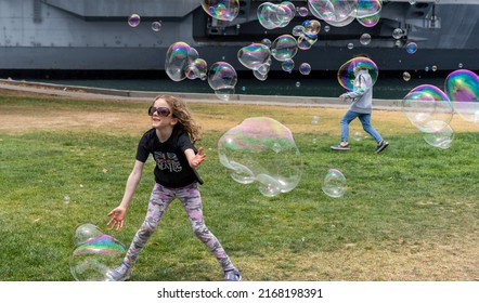 San Diego, California, USA - 5 June 2022: Children Play On Grass In Tuna Harbor Park Chasing Soap Bubbles, Which Shine Irridescent Against The Grey Hull Of USS Midway. They Laugh And Run, Having Fun.