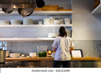 SAN FRANCISCO, CA- April 14, 2017: Restaurant Kitchen Worker In Prep Area Seen From The Back. Open Shelving With Stacks Of Dishes. Overhead Pot Rack.