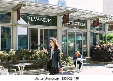 San Francisco, California / United States - November 8 2016: Millennial Businesswoman Holding A Cell Phone And Wearing Ear Buds Walks Past Woman On Laptop Sitting Outside Cafe In Union Square