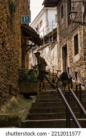 SAN VICENTE DE LA BARQUERA, SPAIN, JULY, 13, 2022: Tourist On Bicycle Entering A Rural Pension To Ask For Accommodation. Travel, Tourism And Vacation Concept.