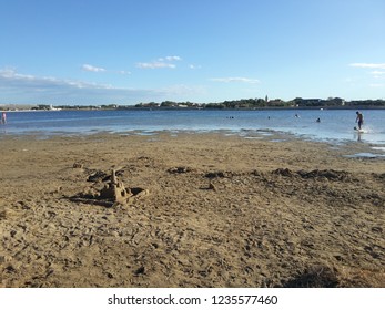 Sand Beach With Sandcastle And City In Distance Behind The Shallow Water, Blue Sky Above