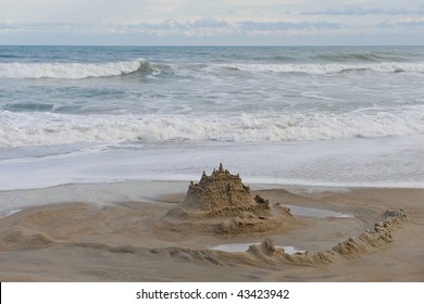 Sand Castle Surrounded By A Moat On The Beach.  Ocean And Cloud Filled Sky In The Background.