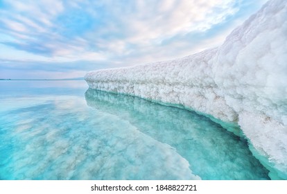 Sand completely covered with crystalline salt looks like ice or snow on shore of Dead Sea, turquoise blue water near, sky colored with morning sun distance - typical scenery at Ein Bokek beach, Israel
