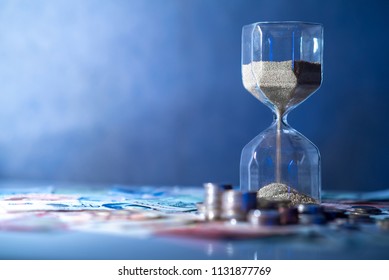 Sand running through the shape of hourglass on table with banknotes and coins of international currency. Time investment and retirement saving. Urgency countdown timer for business deadline concept - Powered by Shutterstock