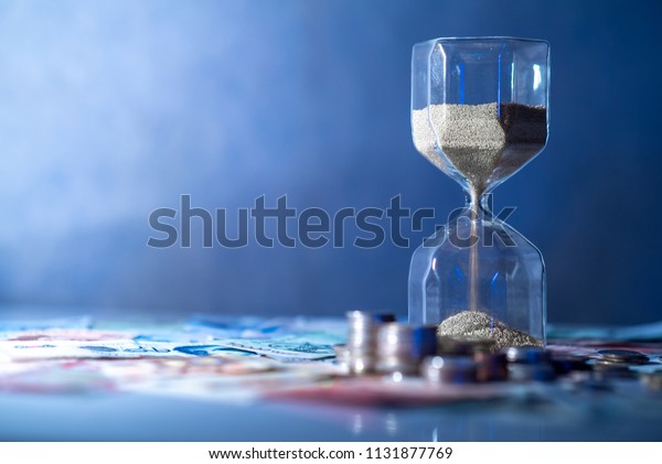 Sand running through the shape of hourglass on\
table with banknotes and coins of international currency. Time\
investment and retirement saving. Urgency countdown timer for\
business deadline concept