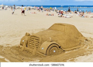 A Sand Sculpture In The Shape Of A Car At La Barceloneta Beach, In Barcelona, Spain, With Unrecognizable People Sunbathing In The Background