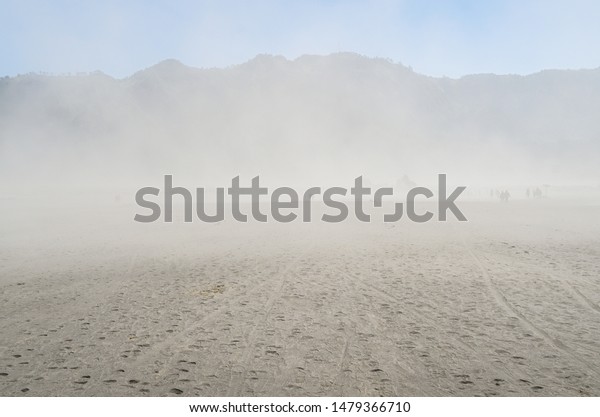Sand storm cover all over a sand\
dune desert with limited vision of mountain and blue\
sky