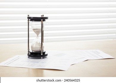 A Sand Timer(hour Glass), Graph Paper(document) On The Wooden Office Desk(table) Behind White Blind.