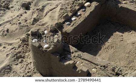 Sandcastle stands alone on empty beach. Walls and towers shaped by childlike imagination. Sunlight highlights textured sand and scattered pebbles. Scene evokes summer, childhood memories