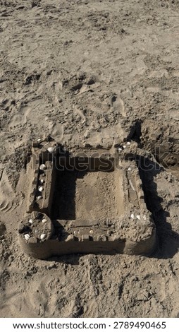 Sandcastle stands alone on empty beach. Walls and towers shaped by childlike imagination. Sunlight highlights textured sand and scattered pebbles. Scene evokes summer, childhood memories