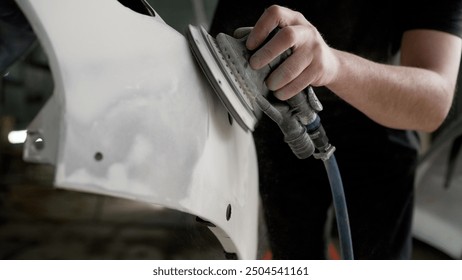 Sanding a car bumper before painting. A worker professionally cleans the surface of a car bumper with a grinding machine in a car service garage before painting, close-up. - Powered by Shutterstock